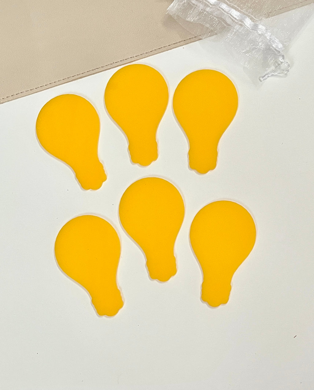 Six bright yellow light bulb-shaped tokens arranged on a white surface next to a sheer drawstring pouch