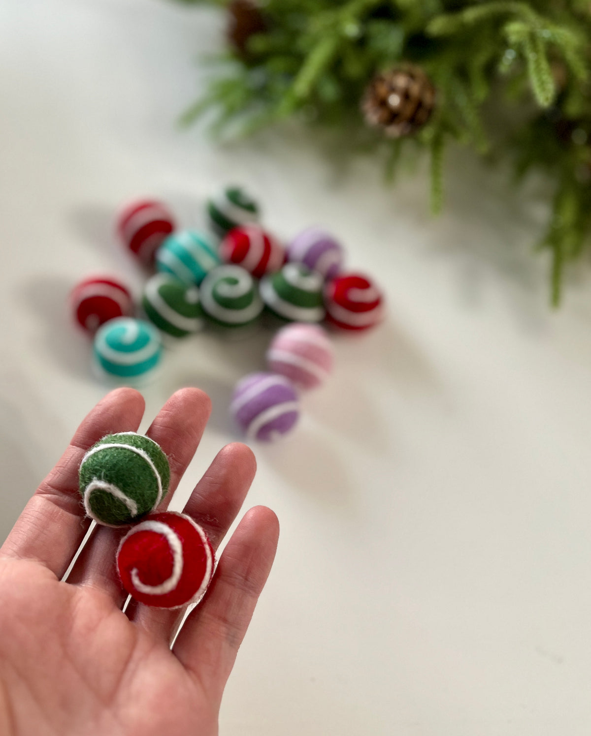Hand holding two felt balls with white swirl patterns in red and green, more colorful felt balls and greenery background visible