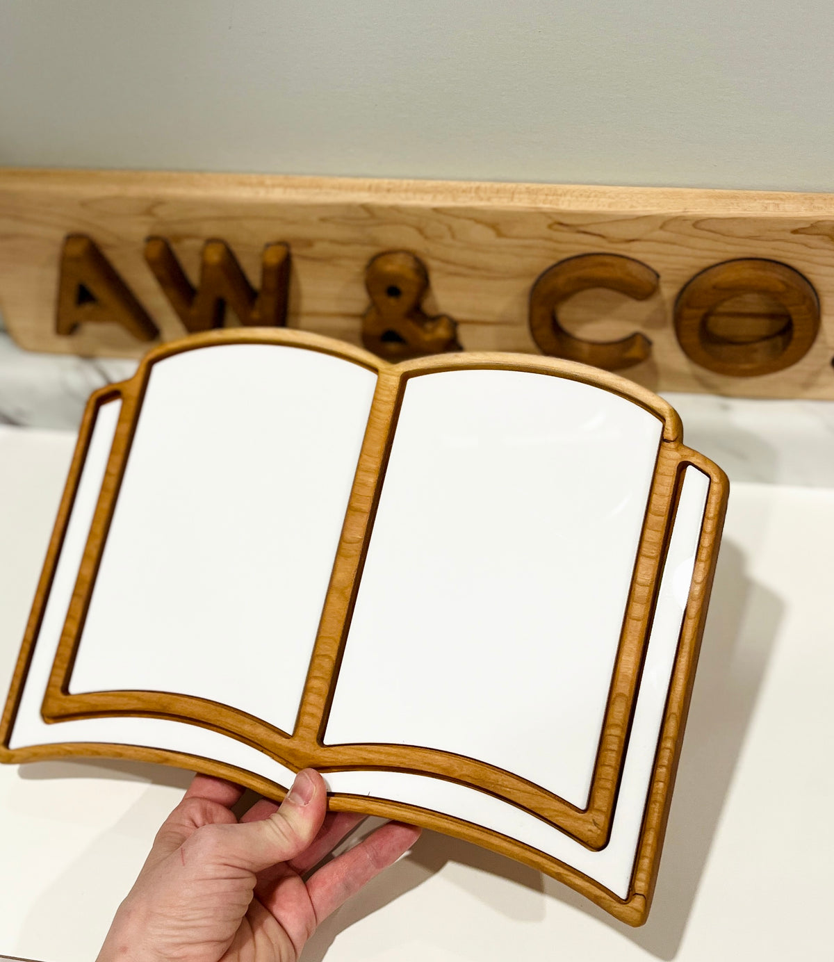 Hand holding an open book-shaped wooden and white ceramic serving tray with a carved wooden sign in background