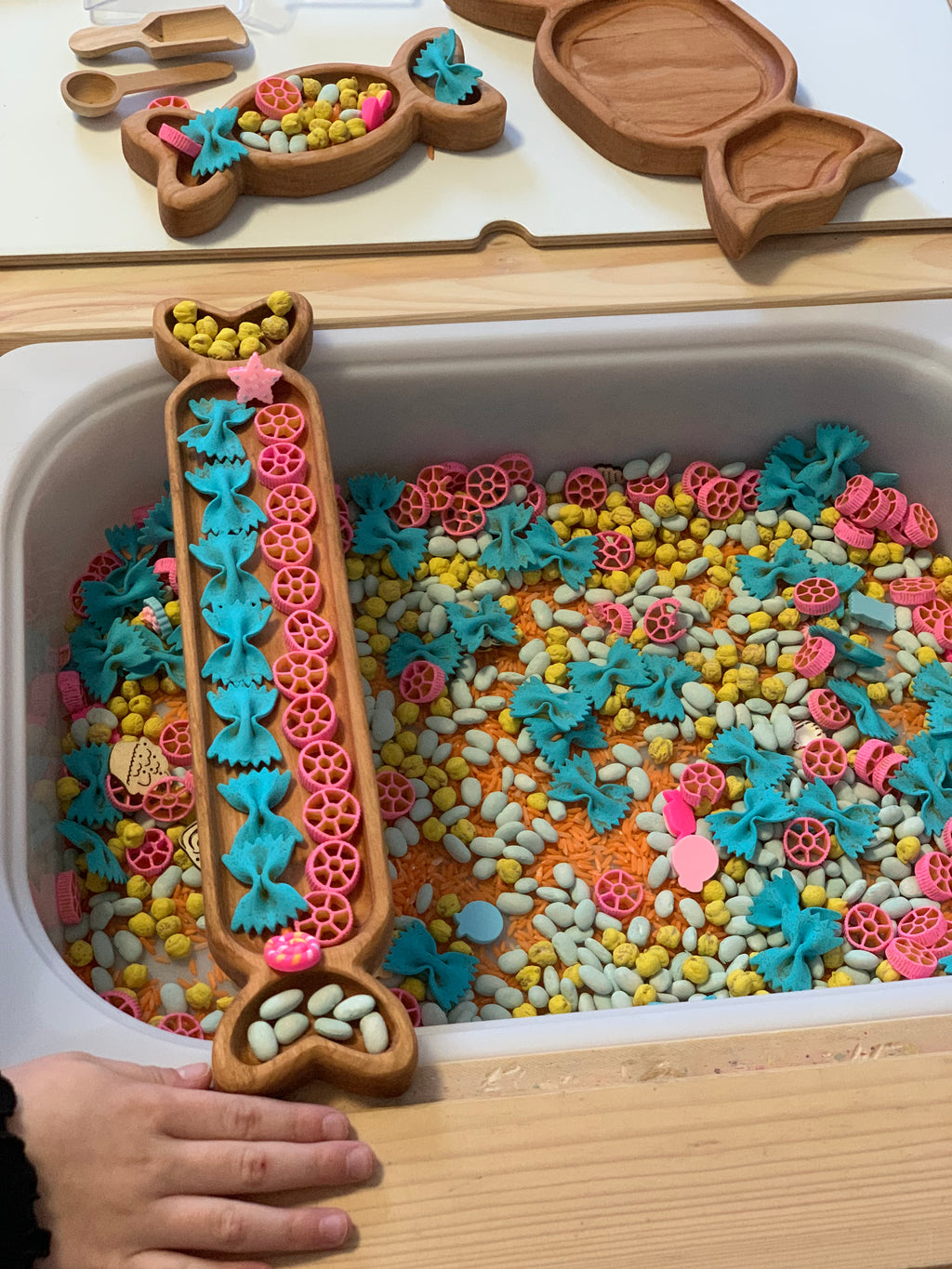 Wooden trays shaped like candies filled with colorful pasta shapes and small wooden tokens in a sensory bin