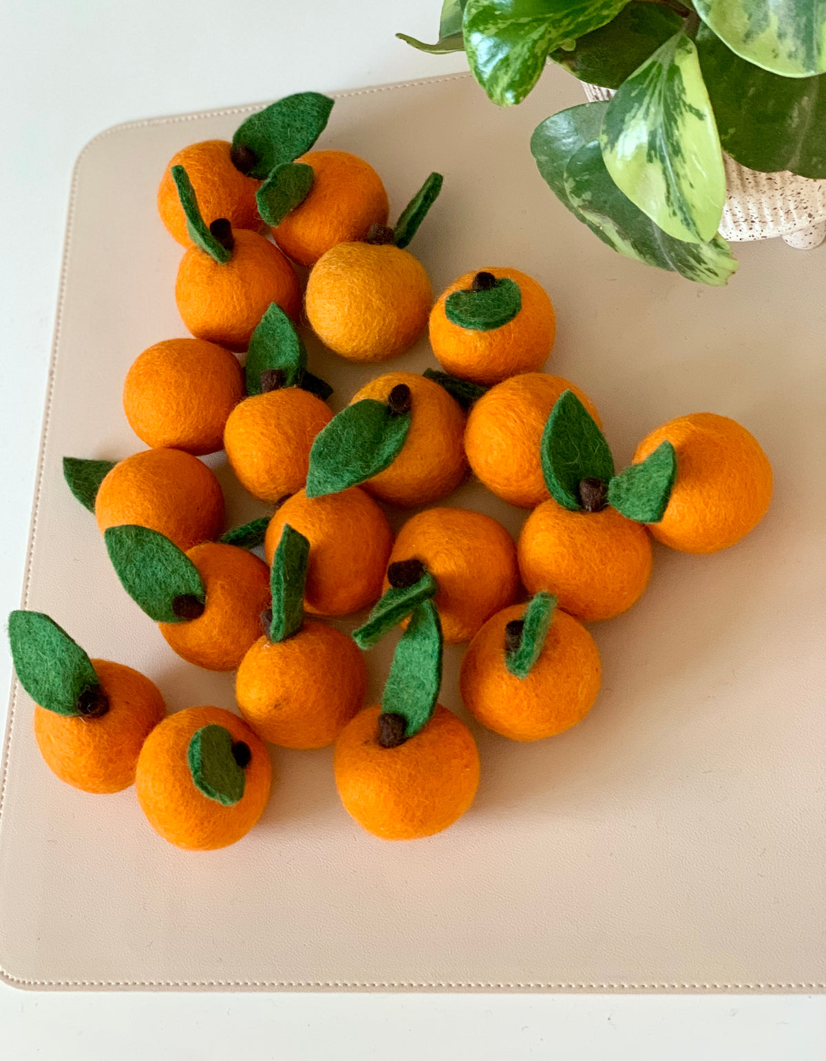 Collection of small felt orange fruits with green leaves arranged on a light tan surface beside a potted plant