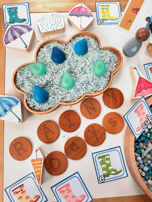 Cloud-shaped sensory tray filled with colored rice and raindrop-shaped manipulatives with wooden letter tiles spelling rainy days