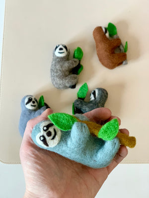 Hand holding a light blue felt sloth figurine clinging to a brown branch with green leaves with four other felt sloths nearby