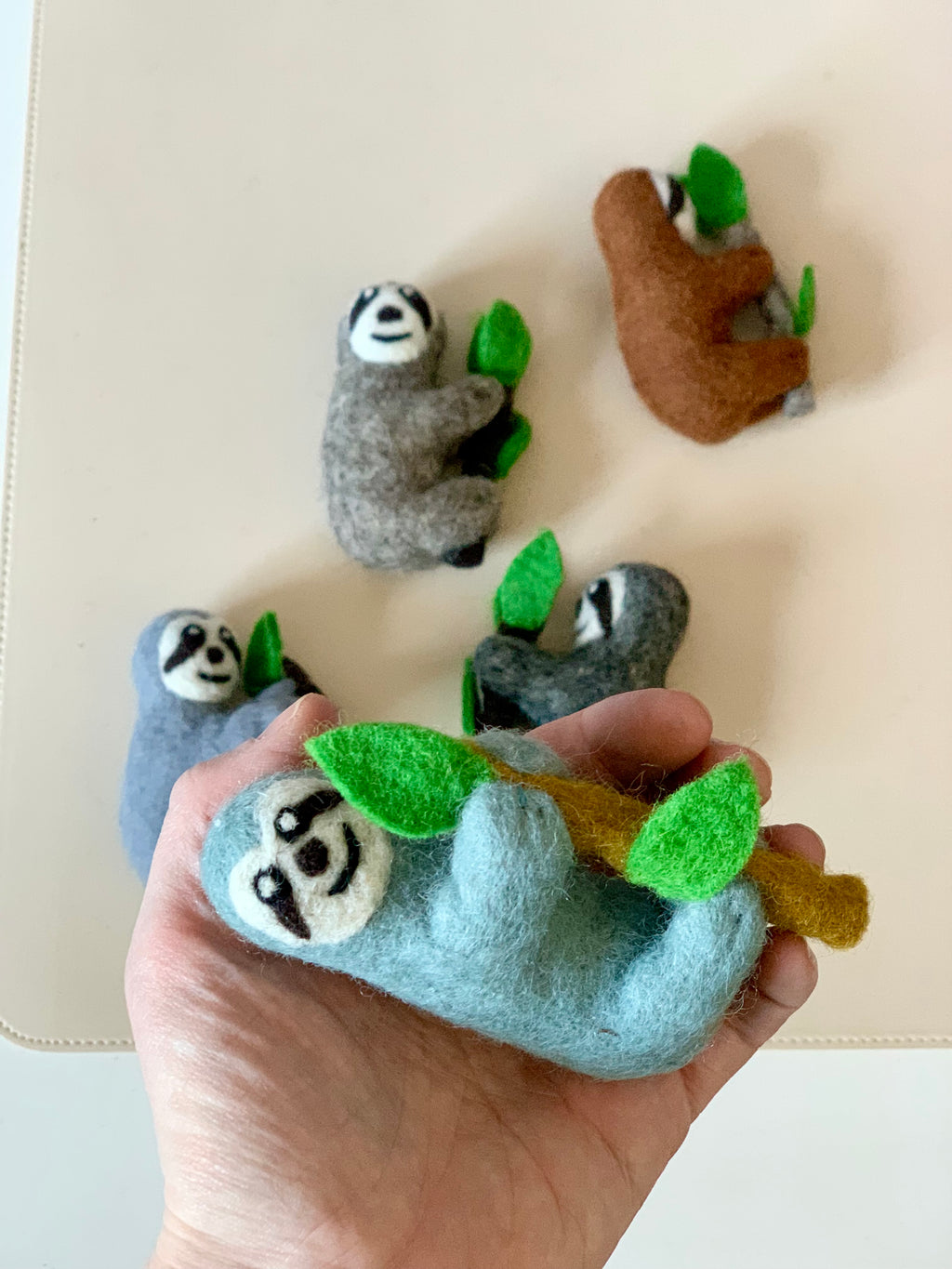 Hand holding a light blue felt sloth figurine clinging to a brown branch with green leaves with four other felt sloths nearby