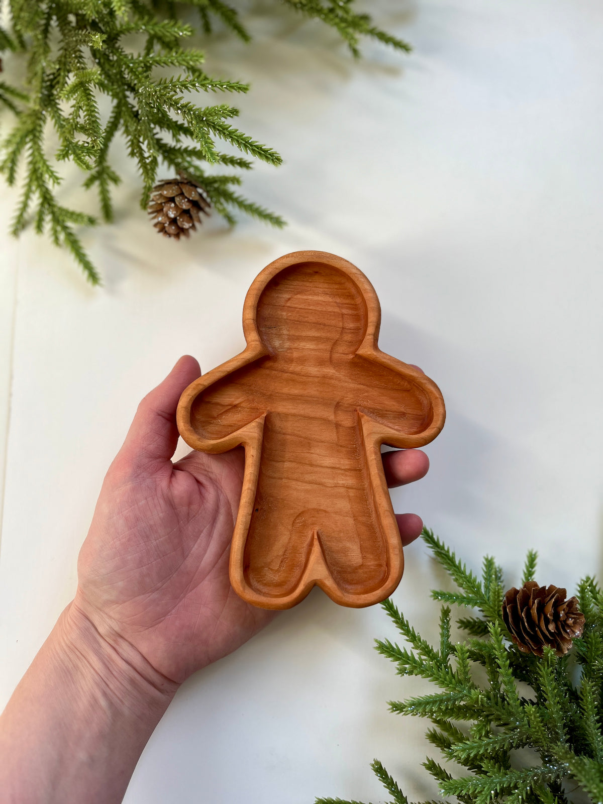 Hand holding a wooden tray shaped like a gingerbread person surrounded by pine branches and pine cones on white surface
