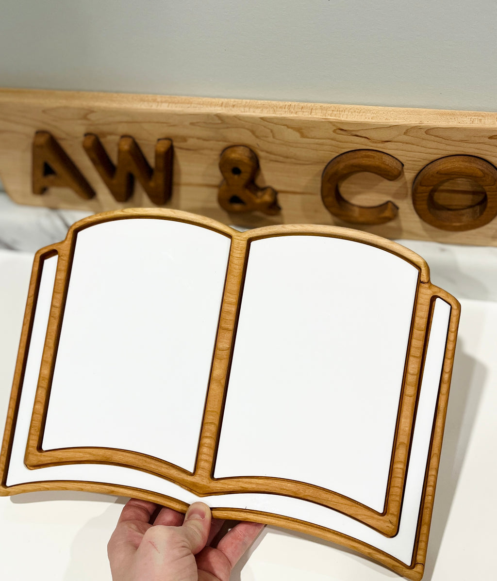 Hand holding a wooden tray shaped like an open book with white inlay on a white surface with wooden letters behind it