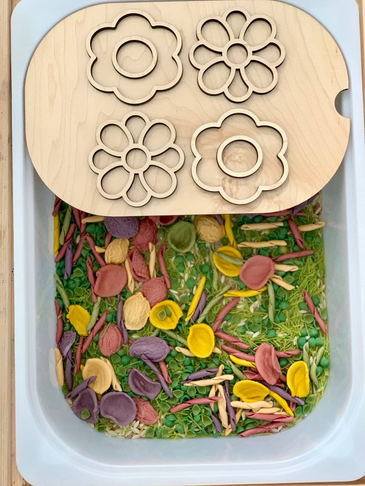 Rectangular white sensory bin filled with colorful dried pasta and green split peas with flower-shaped wooden puzzle lid on top