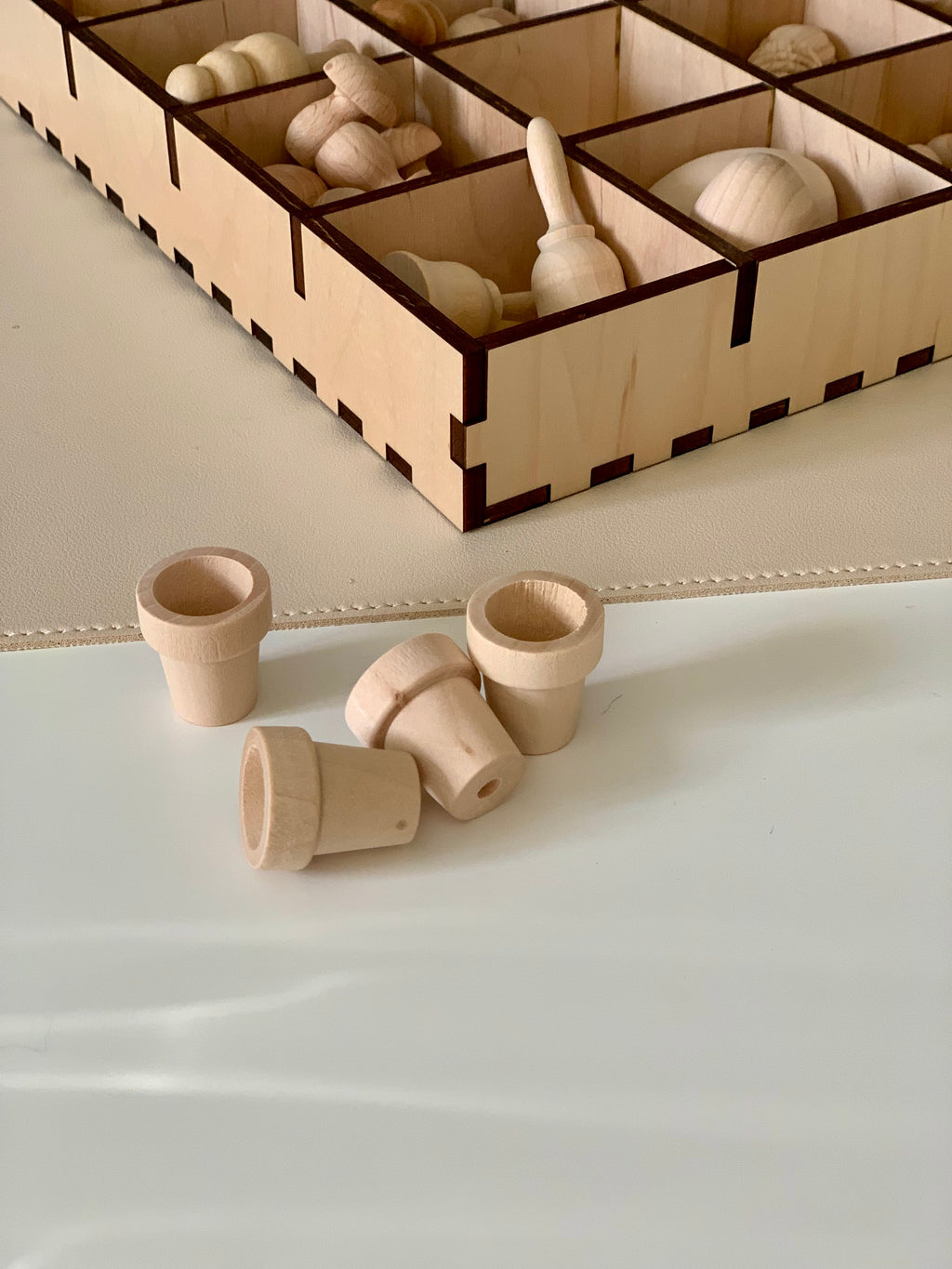 Four small unfinished wooden peg cup shapes on a white surface with a wooden compartment box in the background