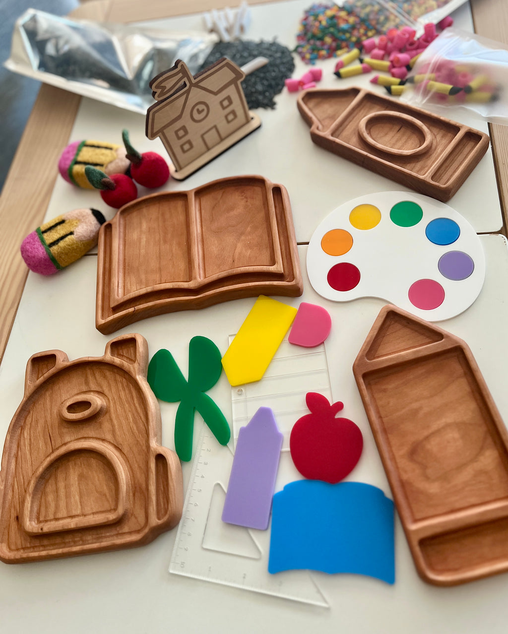 Wooden trays shaped like a book, crayon, house, and backpack surrounded by colorful craft shapes on white surface