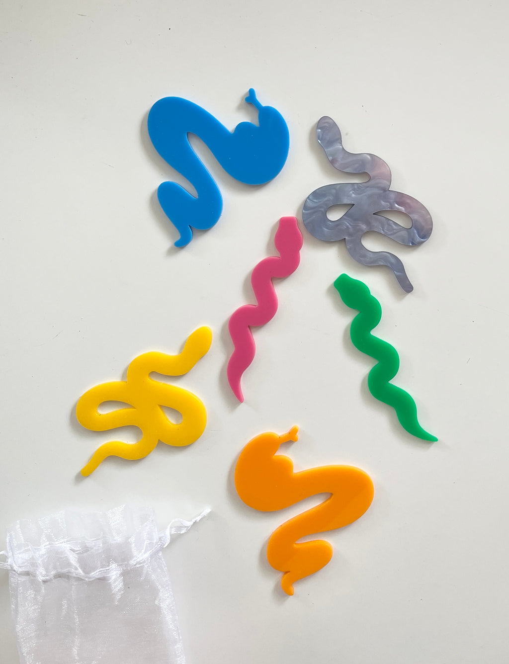 Six colorful snake-shaped cutouts in blue, gray, pink, green, yellow, and orange on a white surface with a sheer white bag nearby