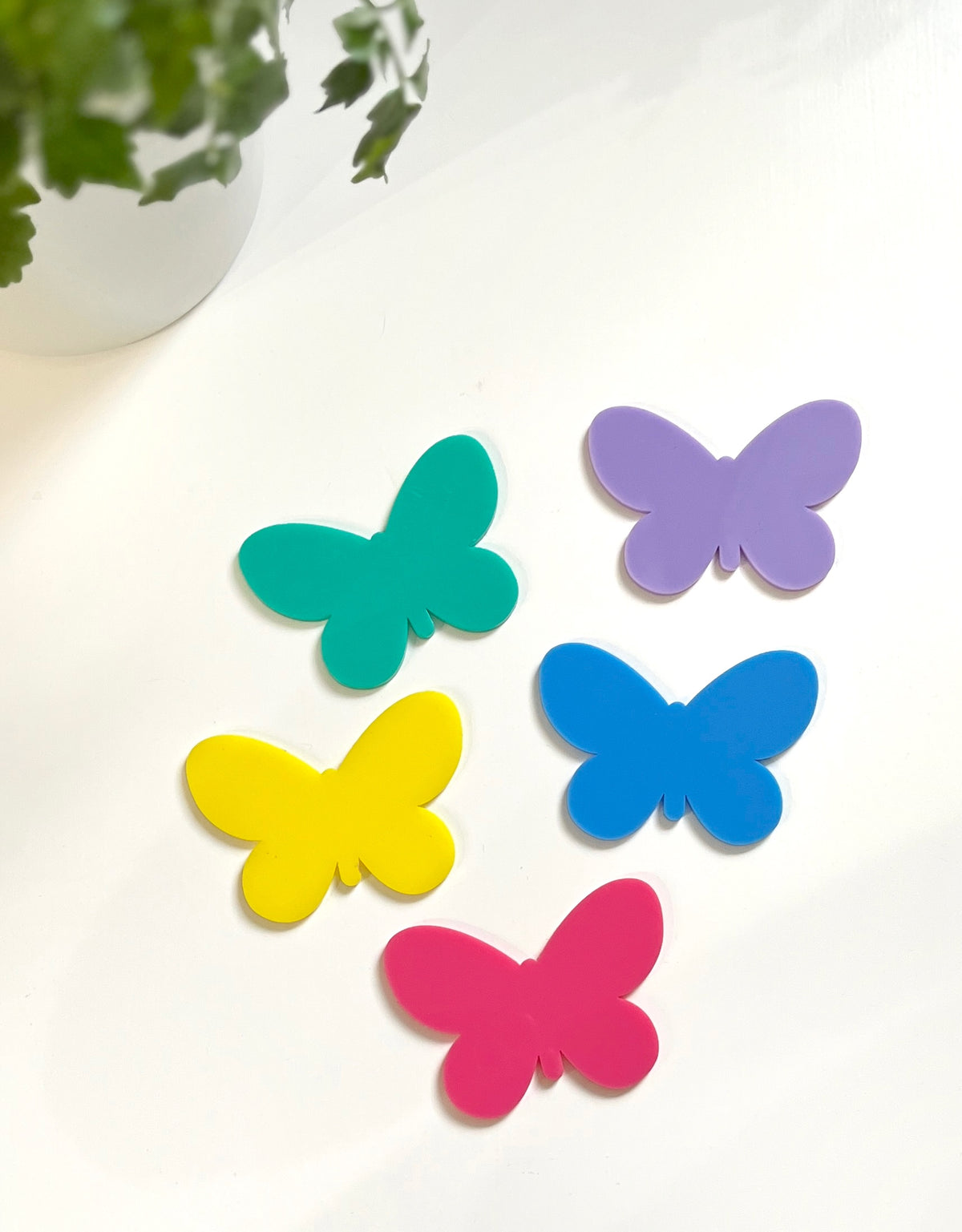 Five colorful butterfly-shaped cutouts in green, purple, yellow, blue, and pink on a white surface near a green plant pot