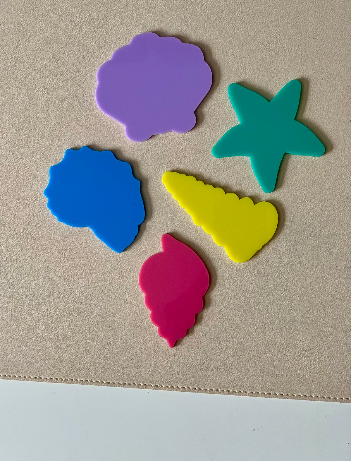 Five colorful plastic shapes including purple scallop shell, teal starfish, blue coral, yellow conch, and pink shell on beige surface