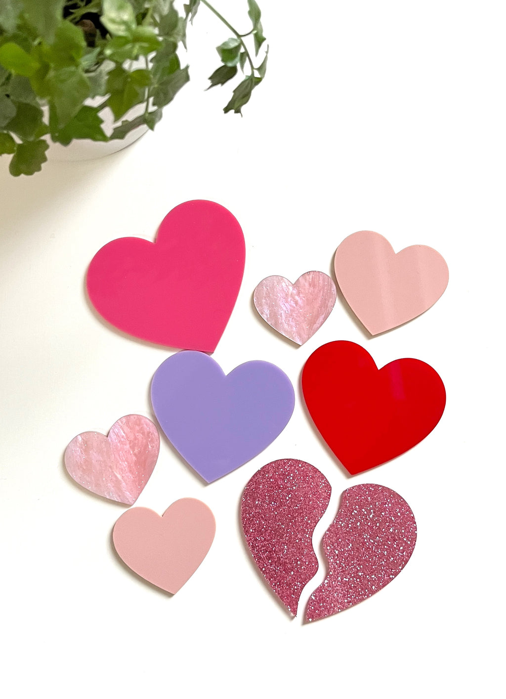 Assortment of flat heart shapes in red, pink, purple, and glittery textures arranged on a white surface near green plant leaves