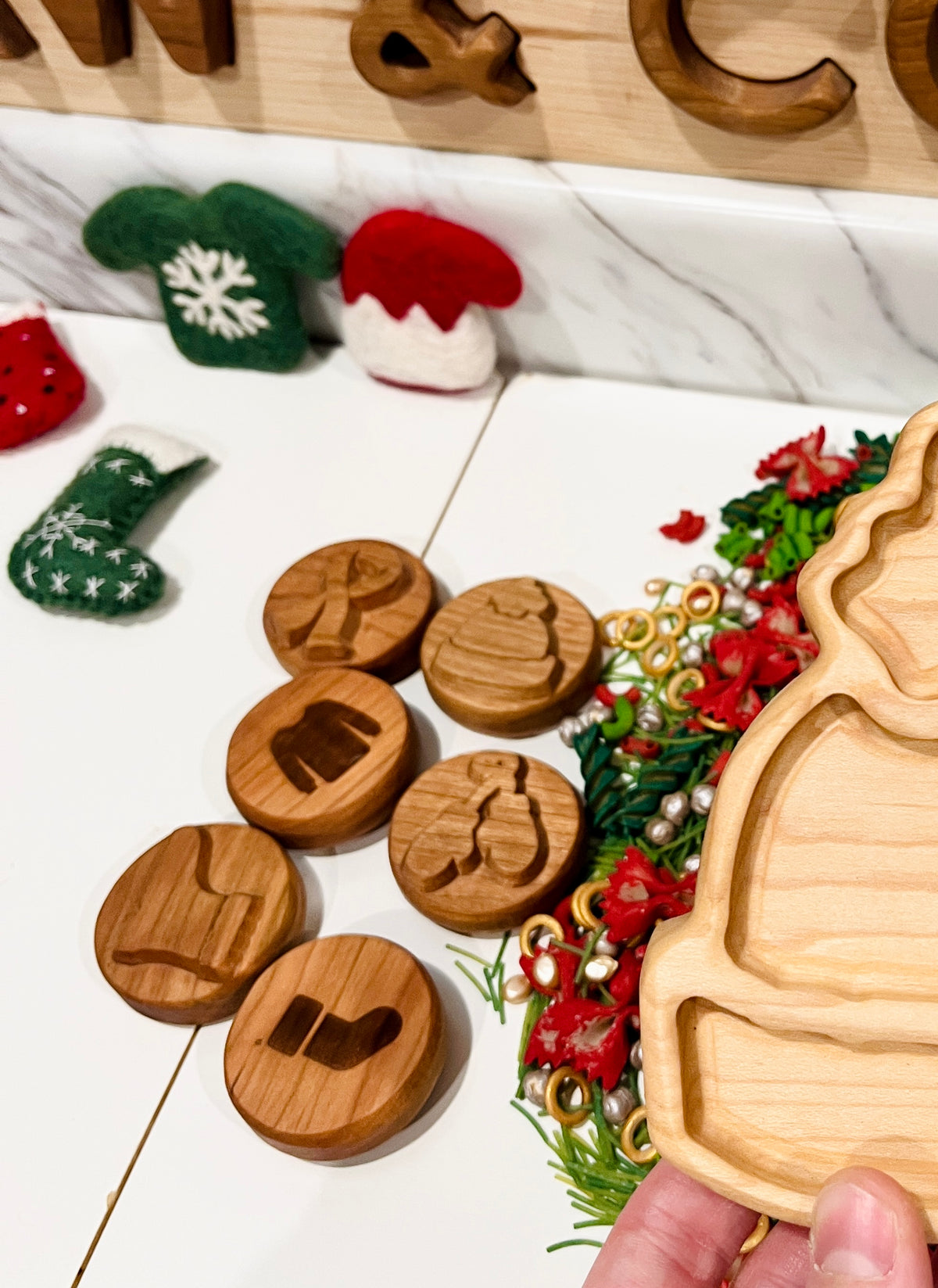 Hand holding wooden Christmas tree tray near round wooden tokens with holiday symbols and felt decorations on white surface