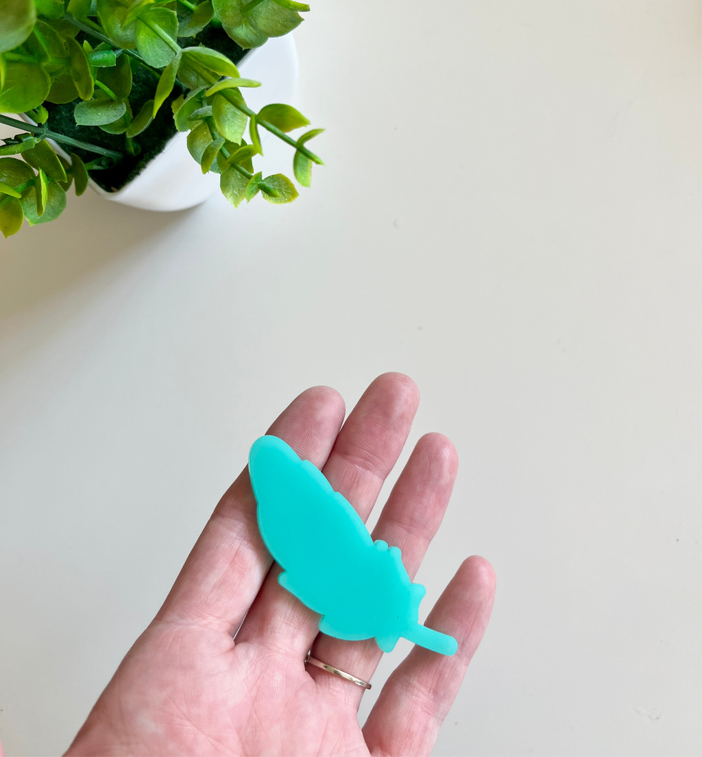 Person holding a smooth turquoise feather-shaped cutout with a green potted plant in the top corner on a light surface