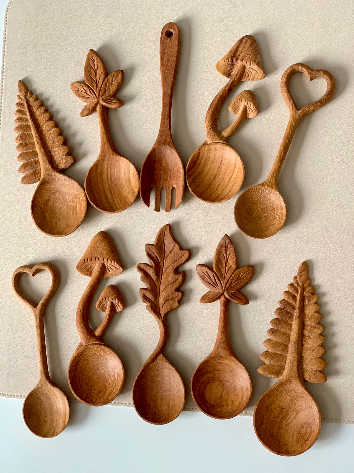 Ten wooden spoons with handles carved into leaves, mushrooms, and heart shapes arranged on a light surface