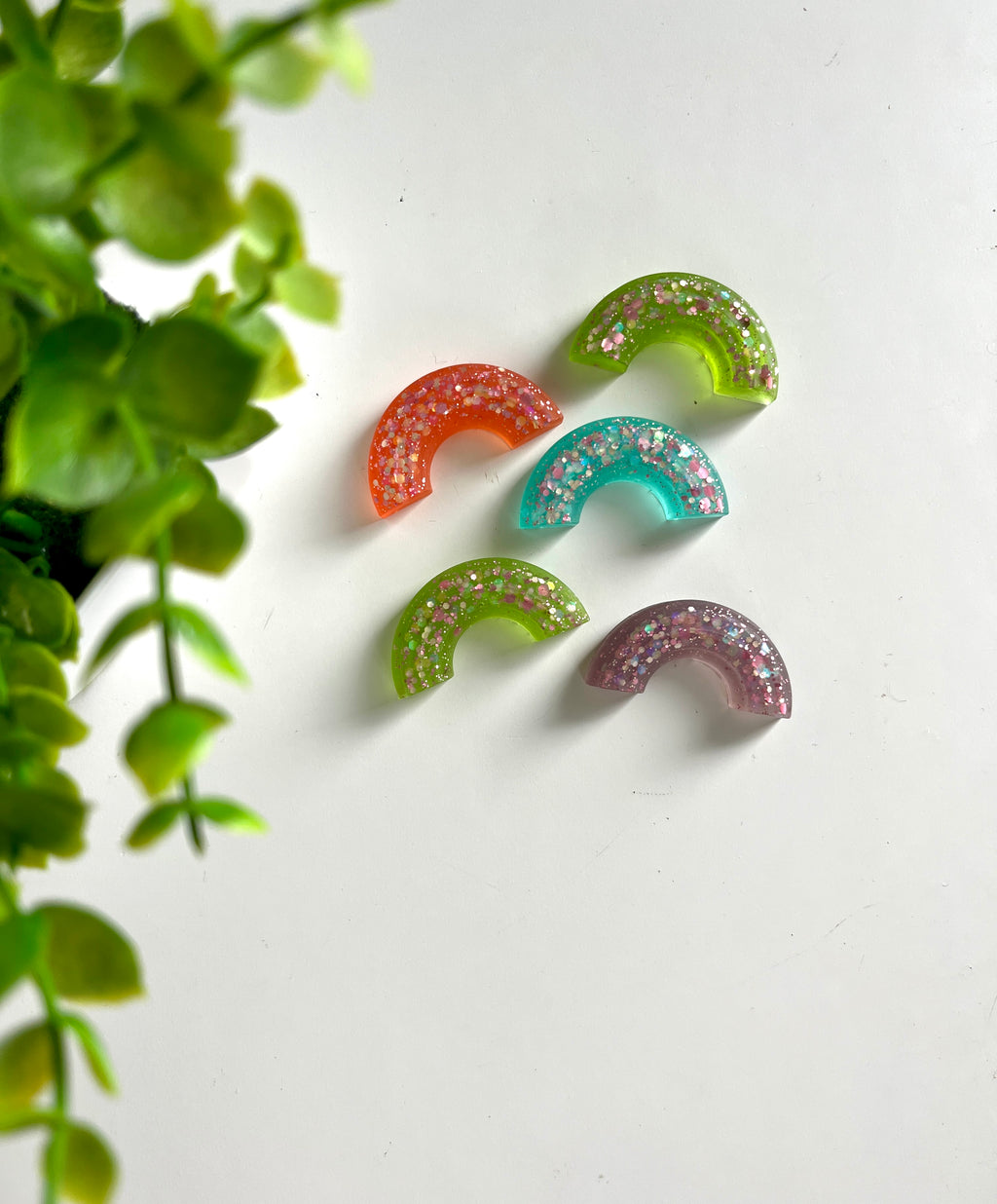 Five small glitter-filled resin pieces in various colors arranged on a white surface near green leaves