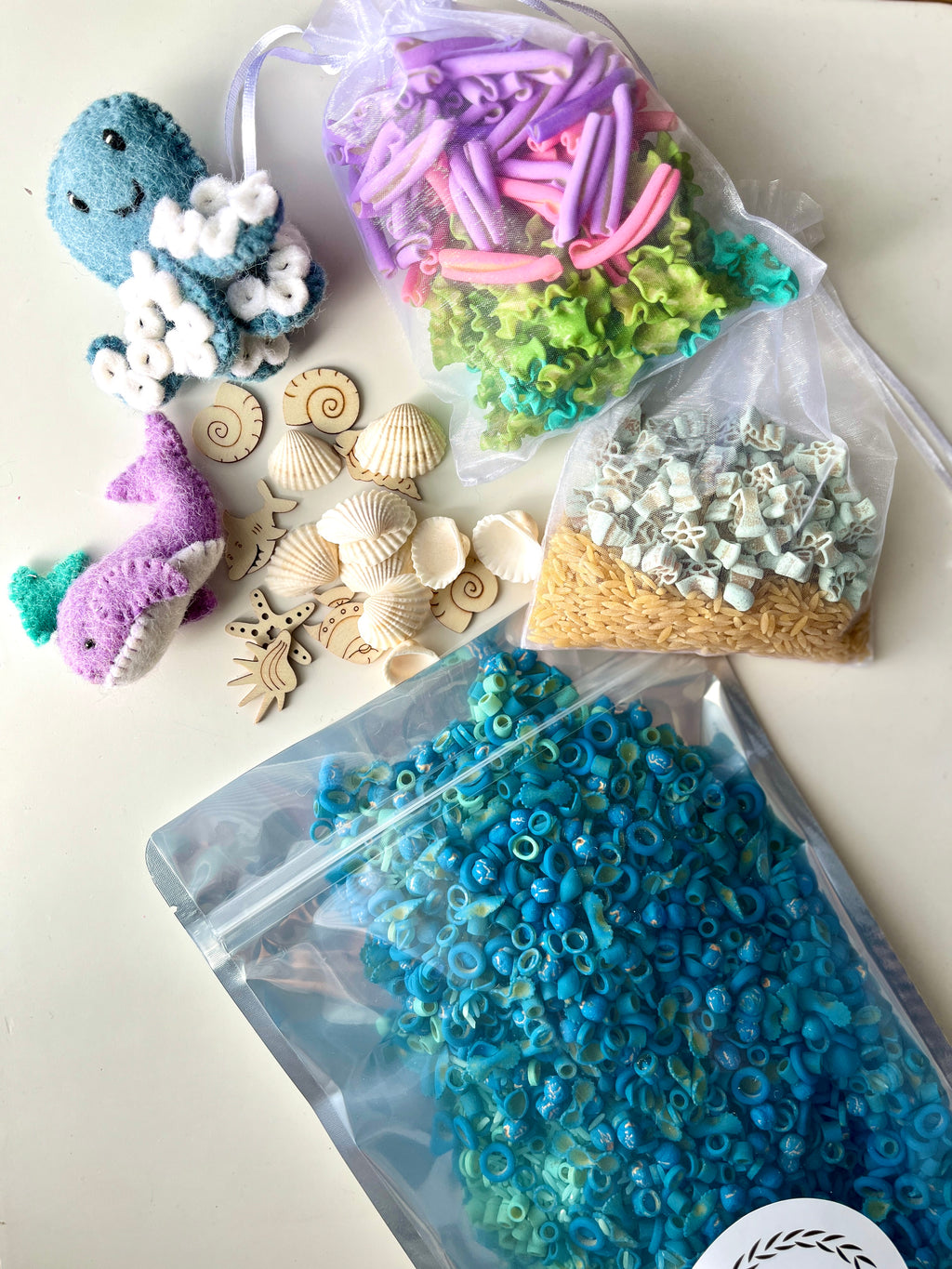 Assorted sensory play items including fabric fish, wooden seashell shapes, colorful pasta, and blue seed beads in resealable bags