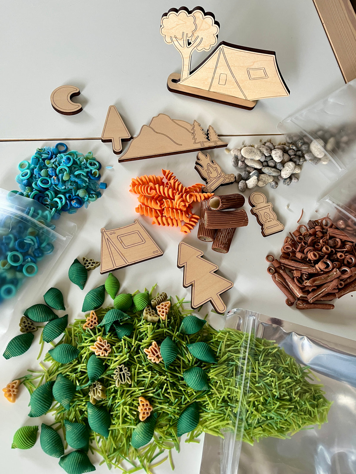 Assorted wooden camping shapes, colorful pasta pieces, and natural textured play items arranged on white surface