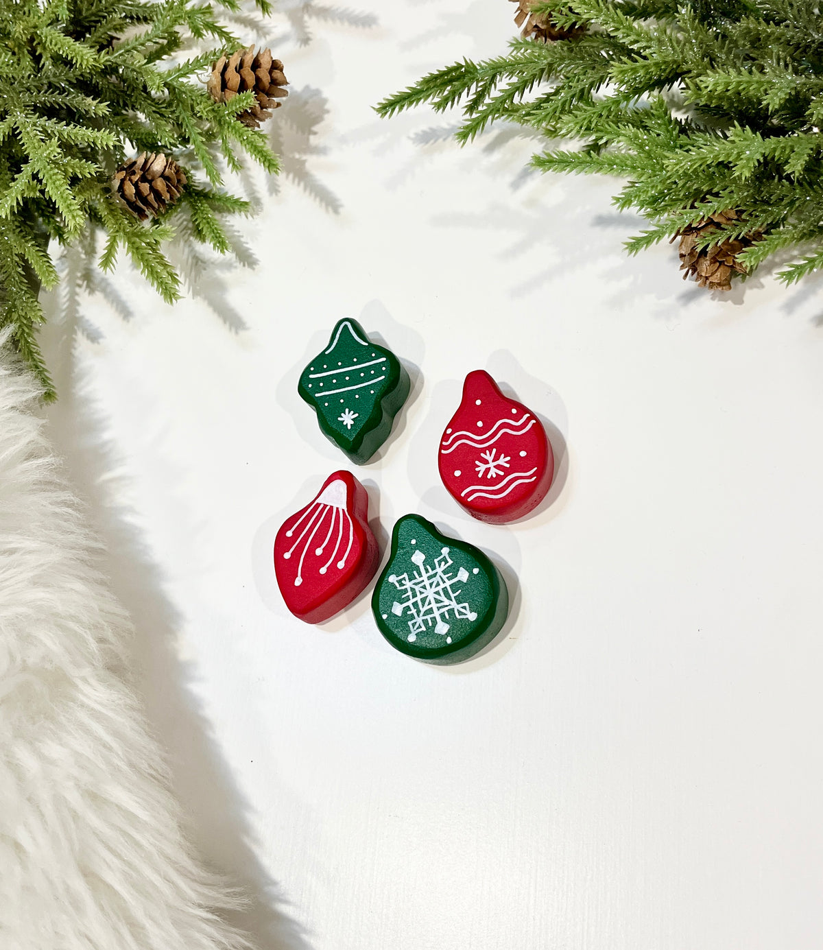 Red and green wooden holiday ornaments with white painted snowflake and stripe designs surrounded by pine branches and cones