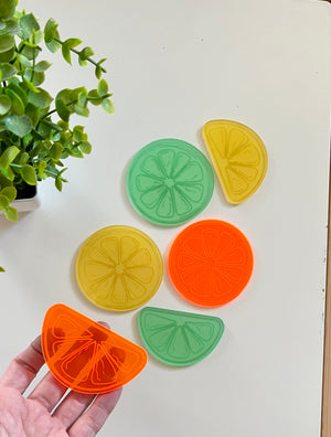 Hand holding an orange translucent citrus slice piece next to yellow and green round and wedge-shaped pieces on white surface