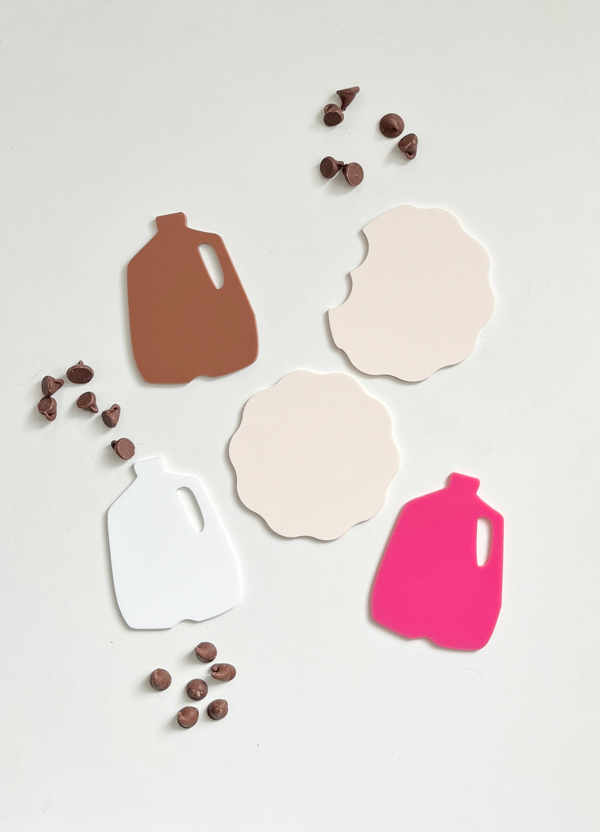 Three jug-shaped cutouts in brown, white, and pink with two scalloped beige shapes and scattered chocolate chips on white surface