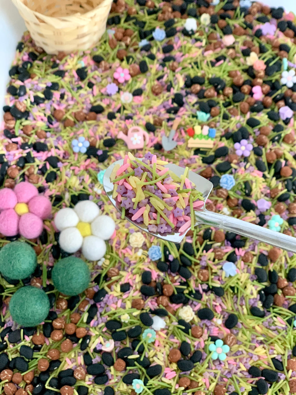 Close-up of colorful rice, small beads, flower-shaped sprinkles, and green pasta pieces with a wicker basket in the background