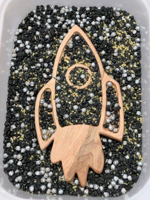 Wooden rocket-shaped tray filled with black, silver, and gold sensory beads inside a white container