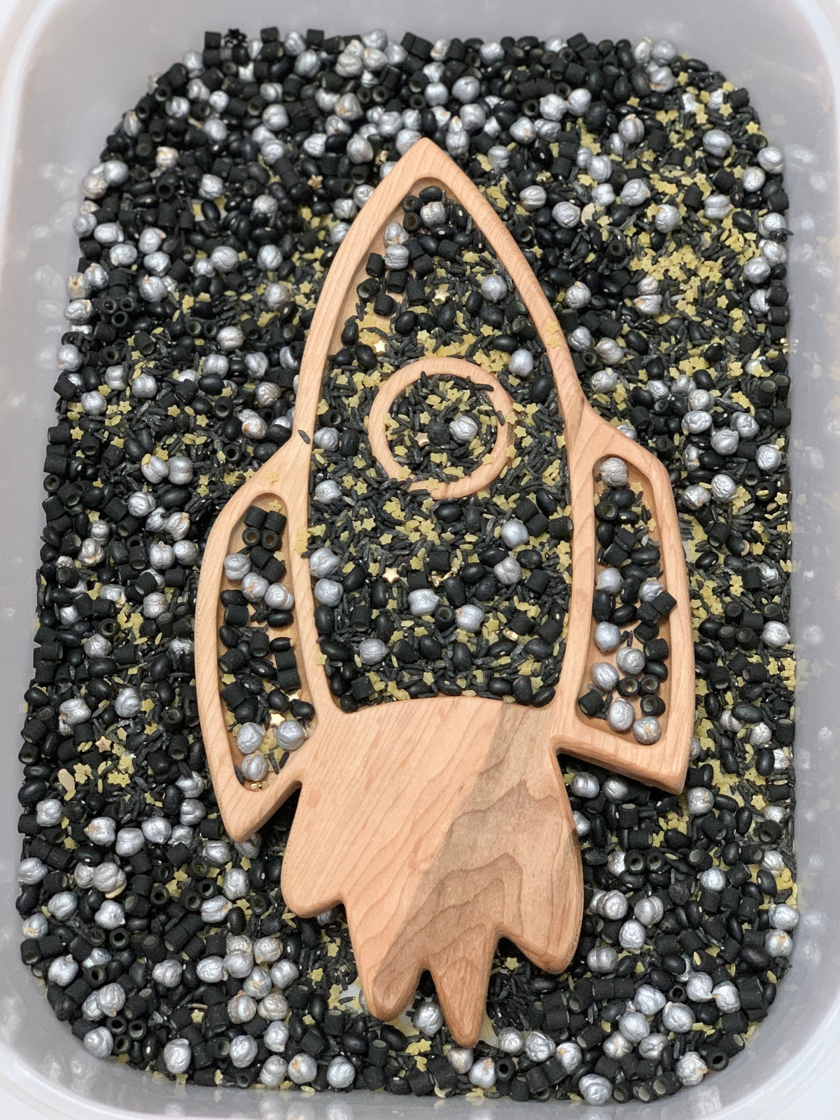 Wooden rocket-shaped tray filled with black, silver, and gold sensory beads inside a white container