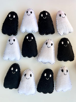 Twelve small handmade felt ghosts in black and white with stitched edges and smiling faces on a light surface
