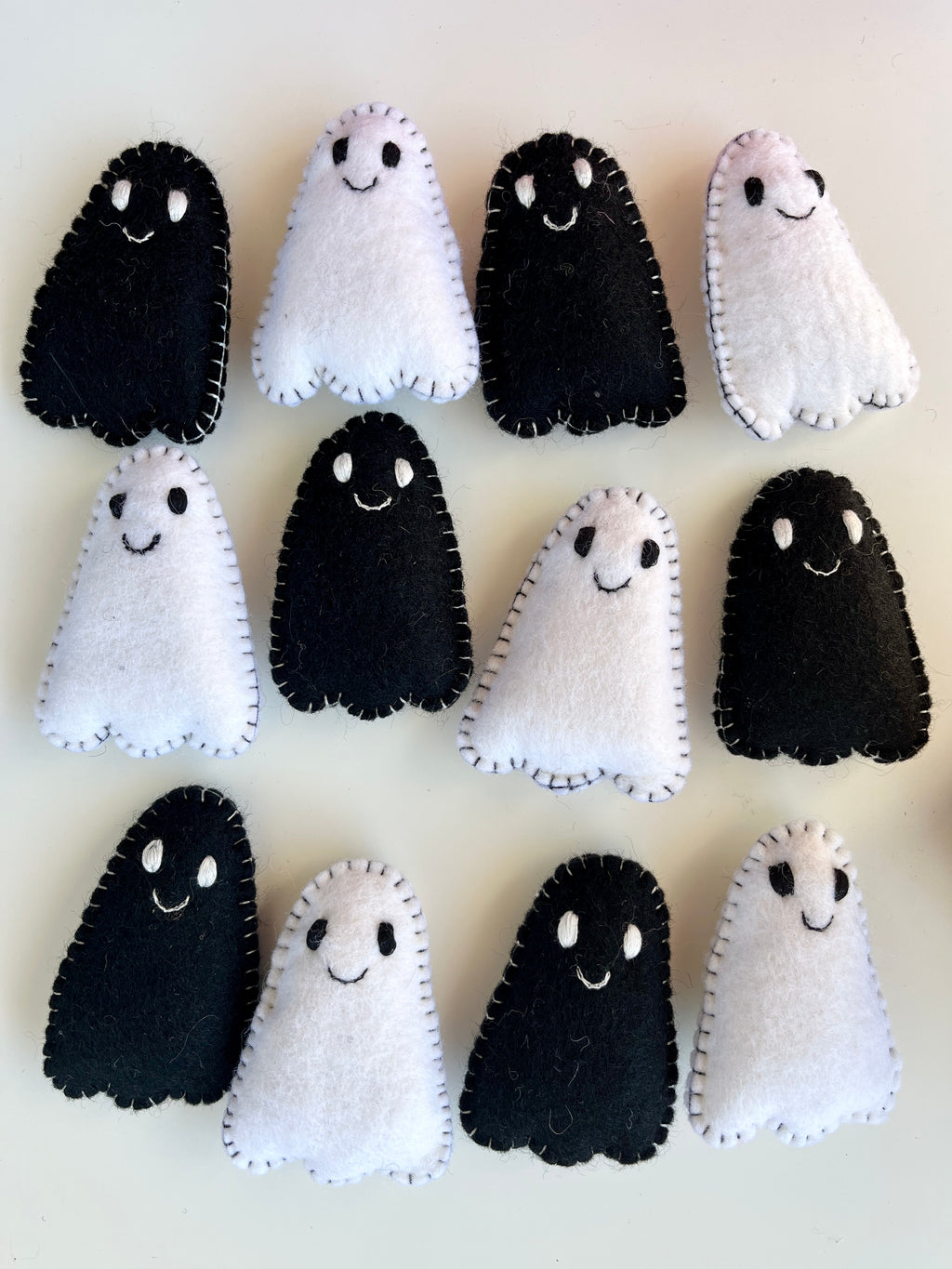 Twelve small handmade felt ghosts in black and white with stitched edges and smiling faces on a light surface