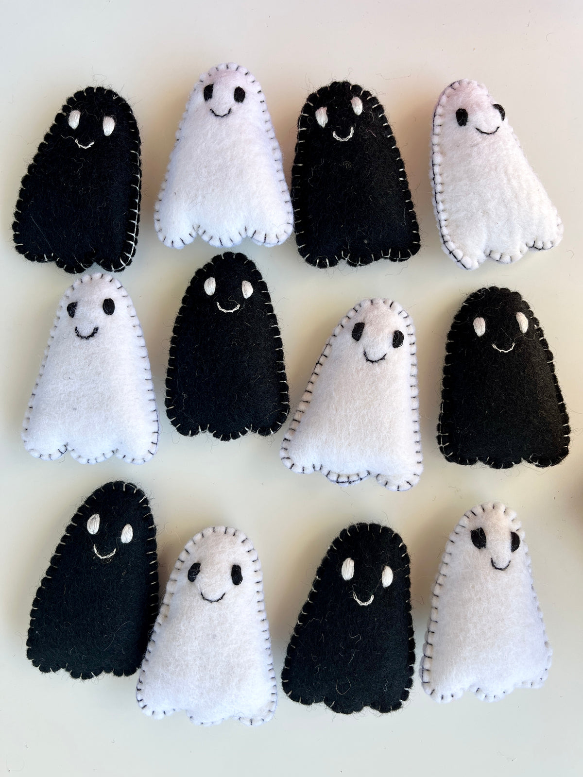 Twelve small handmade felt ghosts in black and white with stitched edges and smiling faces on a light surface