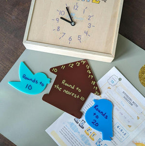 Wooden clock with loose hands, brown house-shaped piece with numbers, and blue bird-shaped pieces labeled with rounding instructions