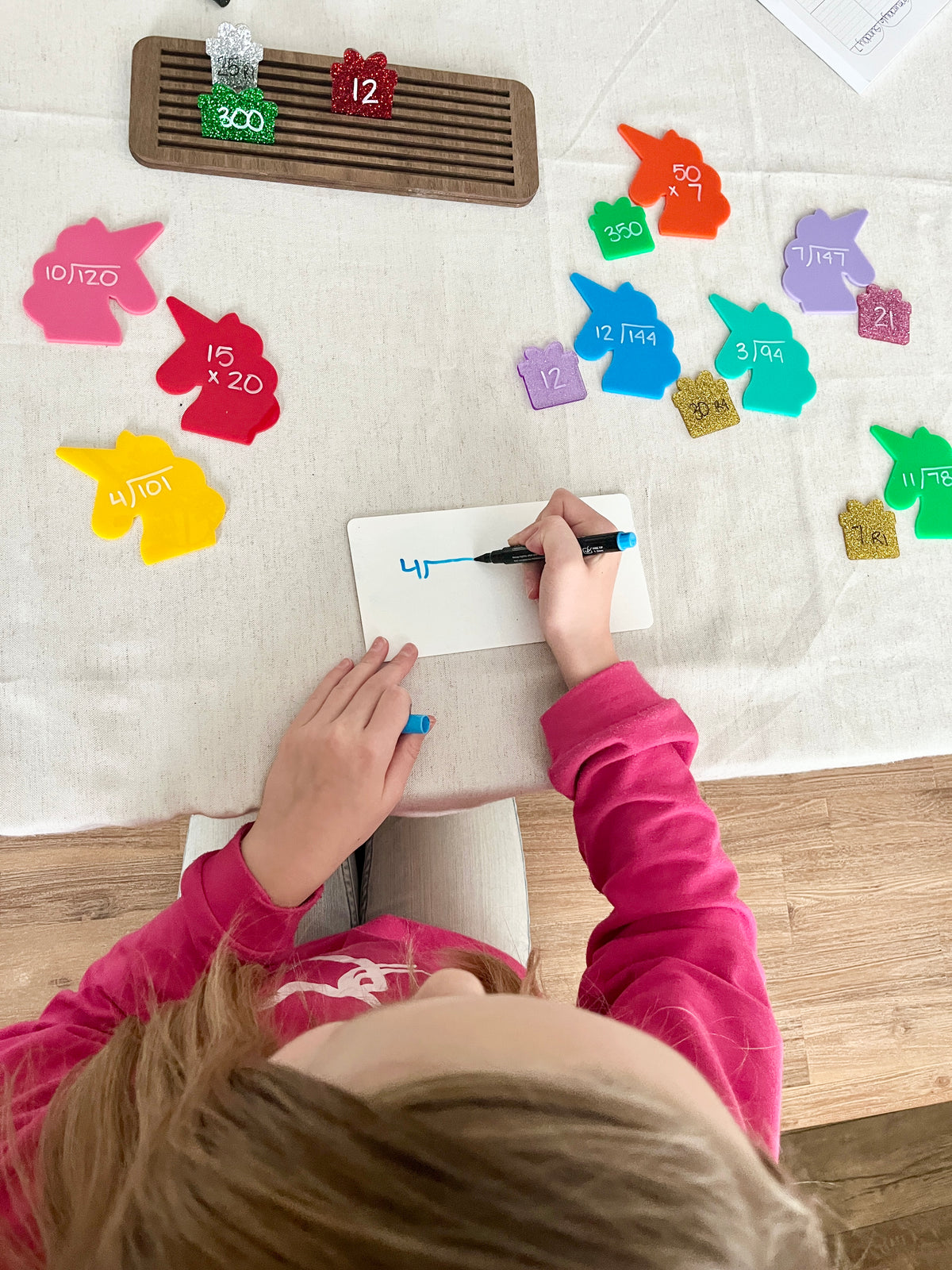Child writing math multiplication problems with a blue marker on a small whiteboard, surrounded by colorful glittery wooden puzzle pieces shaped like animals and gift boxes with handwritten numbers, arranged on a light cloth surface with a wooden sorter tray nearby.