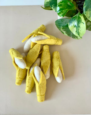 Small handcrafted felt yellow corn cobs with white kernels arranged on a beige surface near green variegated leaves