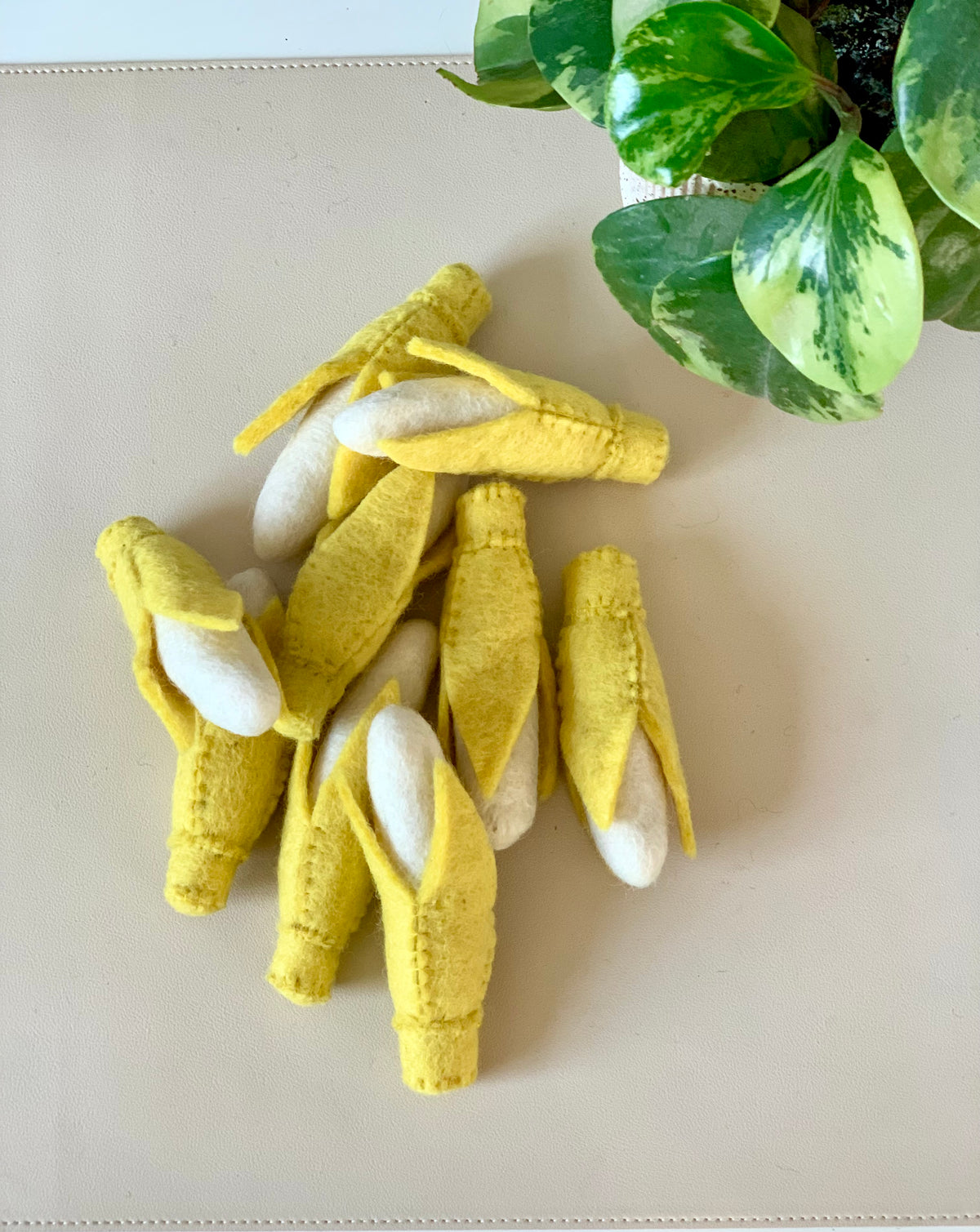 Small handcrafted felt yellow corn cobs with white kernels arranged on a beige surface near green variegated leaves