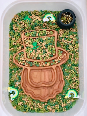 Plastic bin filled with green and beige pasta shapes, wooden leprechaun tray, rainbows, and a small black pot with gold stars
