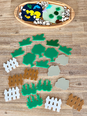 Multipurpose Oval Sensory Tray with natural wooden frame containing blue and translucent green textured sensory fillers, smooth stones, and colorful grass, tree, and fence shapes, handcrafted homeschool education resources.