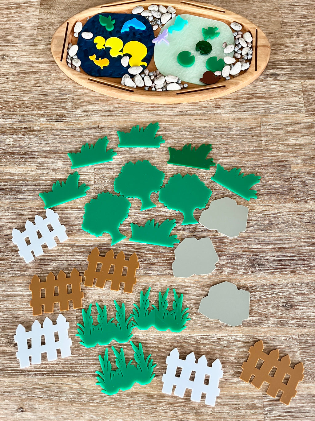 Multipurpose Oval Sensory Tray with natural wooden frame containing blue and translucent green textured sensory fillers, smooth stones, and colorful grass, tree, and fence shapes, handcrafted homeschool education resources.
