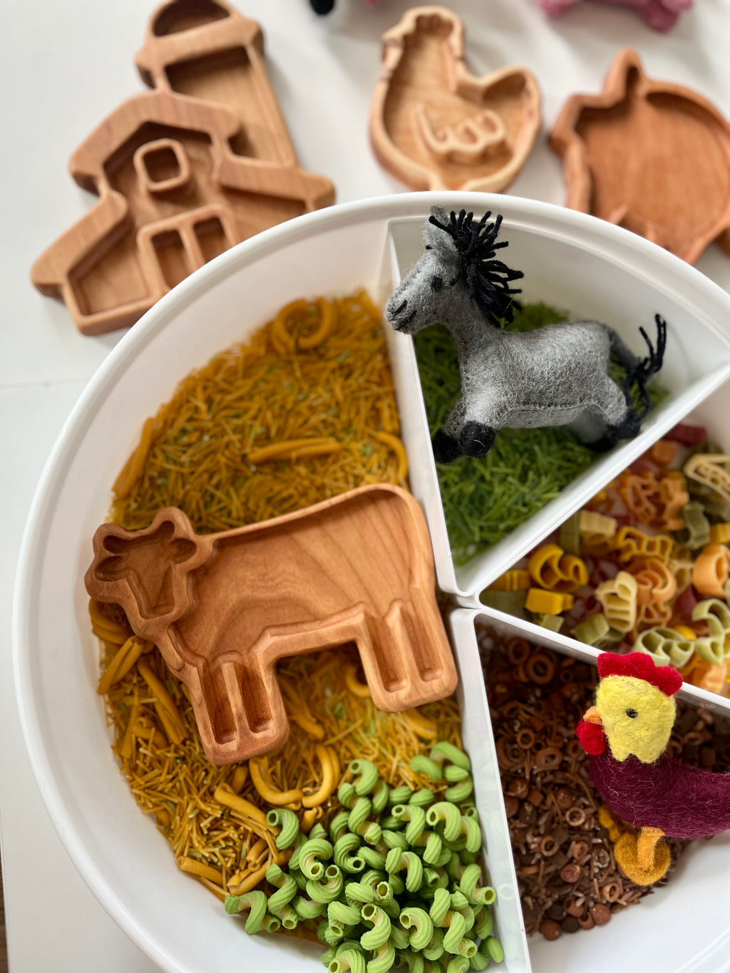 White divided plate with green, yellow, and brown pasta, felt farm animals, and wooden farm-shaped trays on white surface
