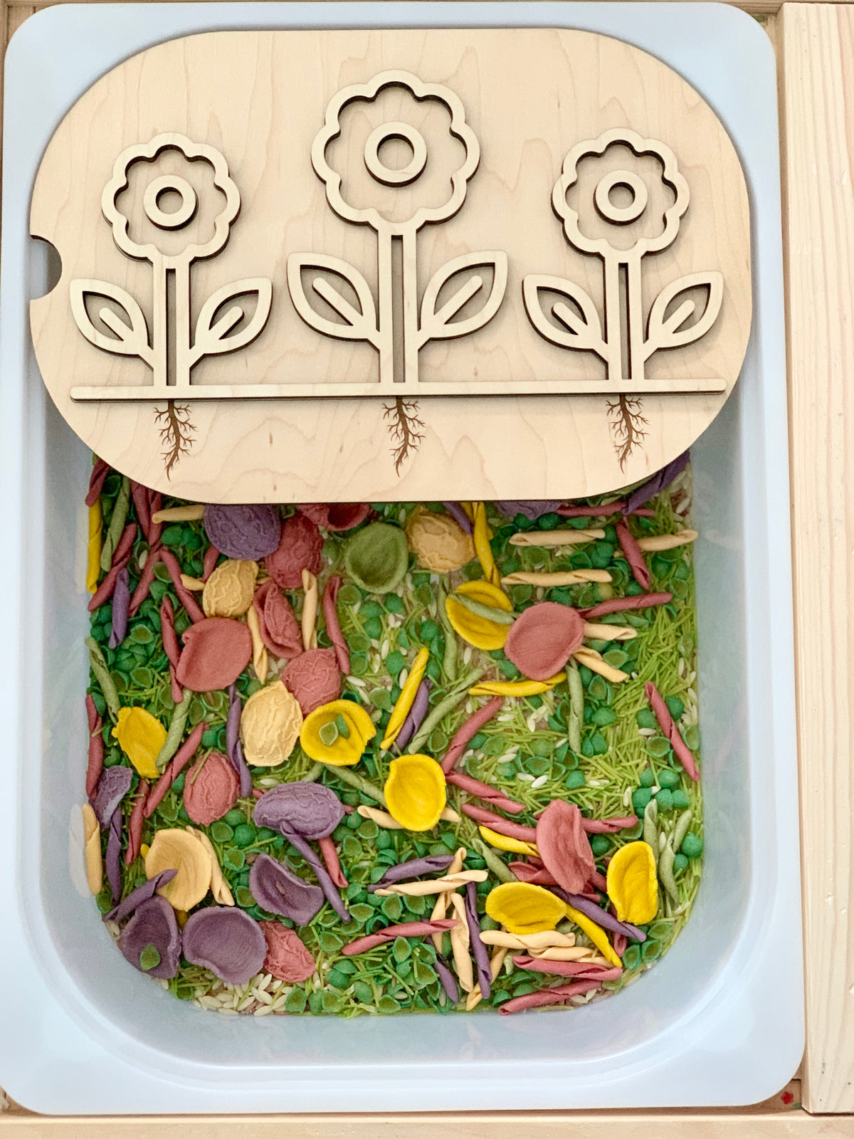 White rectangular sensory bin filled with colorful dried pasta and flower-themed wooden puzzle lid on top