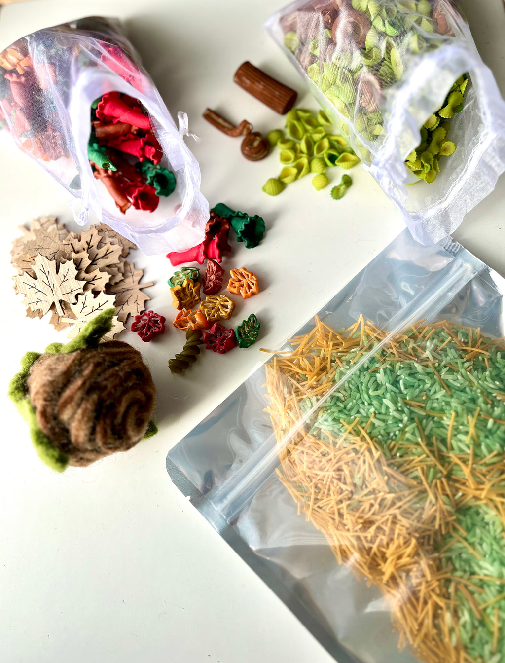 Clear bags spilling green, red, and brown pasta shapes, wooden leaf cutouts, felt pinecone, and mixed dried pasta on white surface