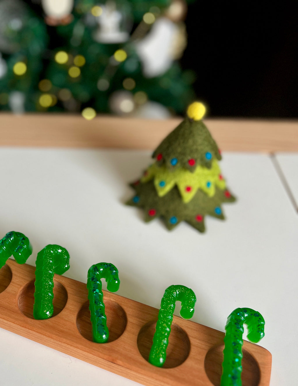 Wooden tray holding green candy cane-shaped objects with blue speckles next to a felt Christmas tree with colorful pom-pom ornaments