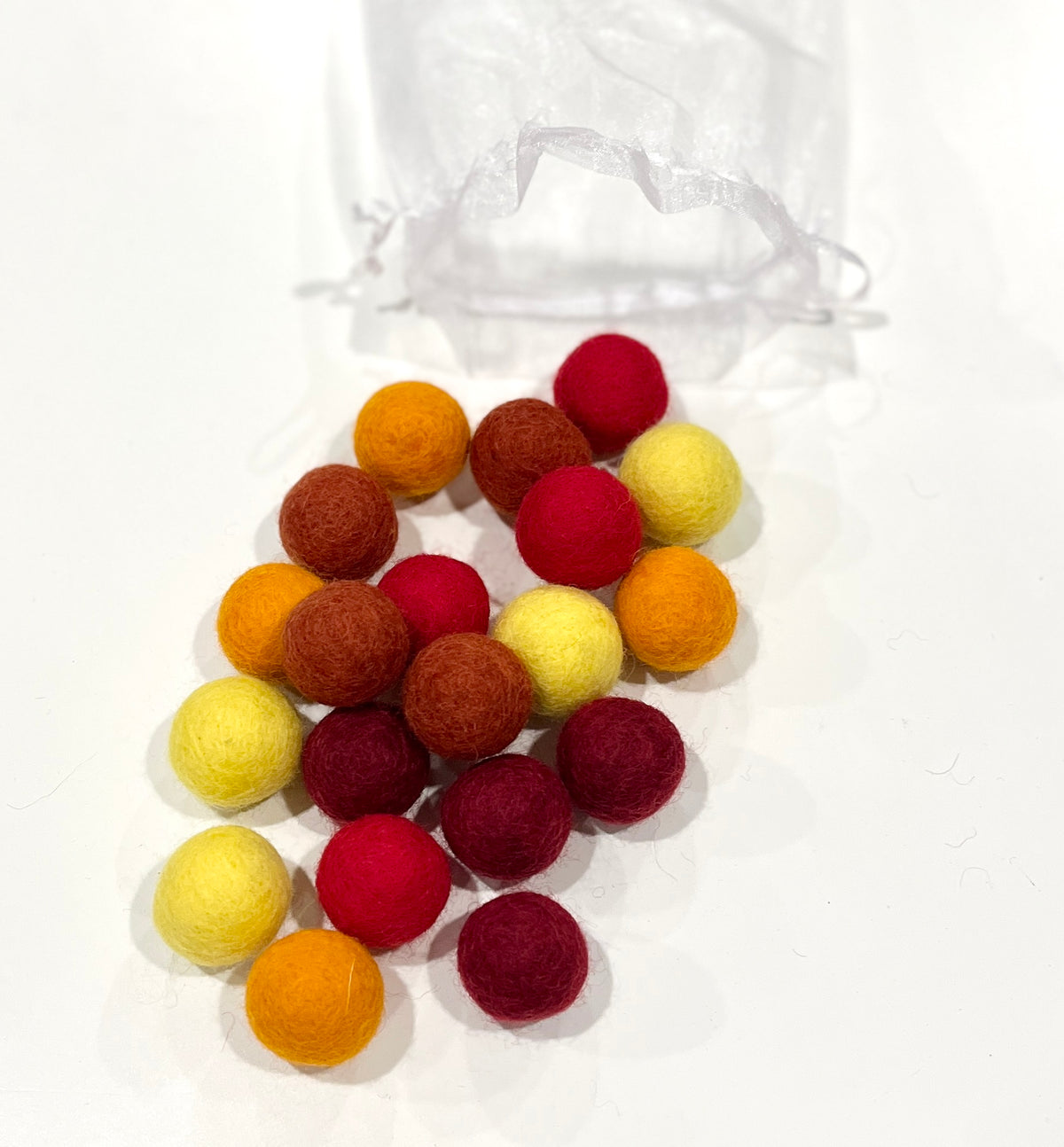 Small round felt balls in red, yellow, orange, and brown shades scattered on a white surface with a sheer bag behind