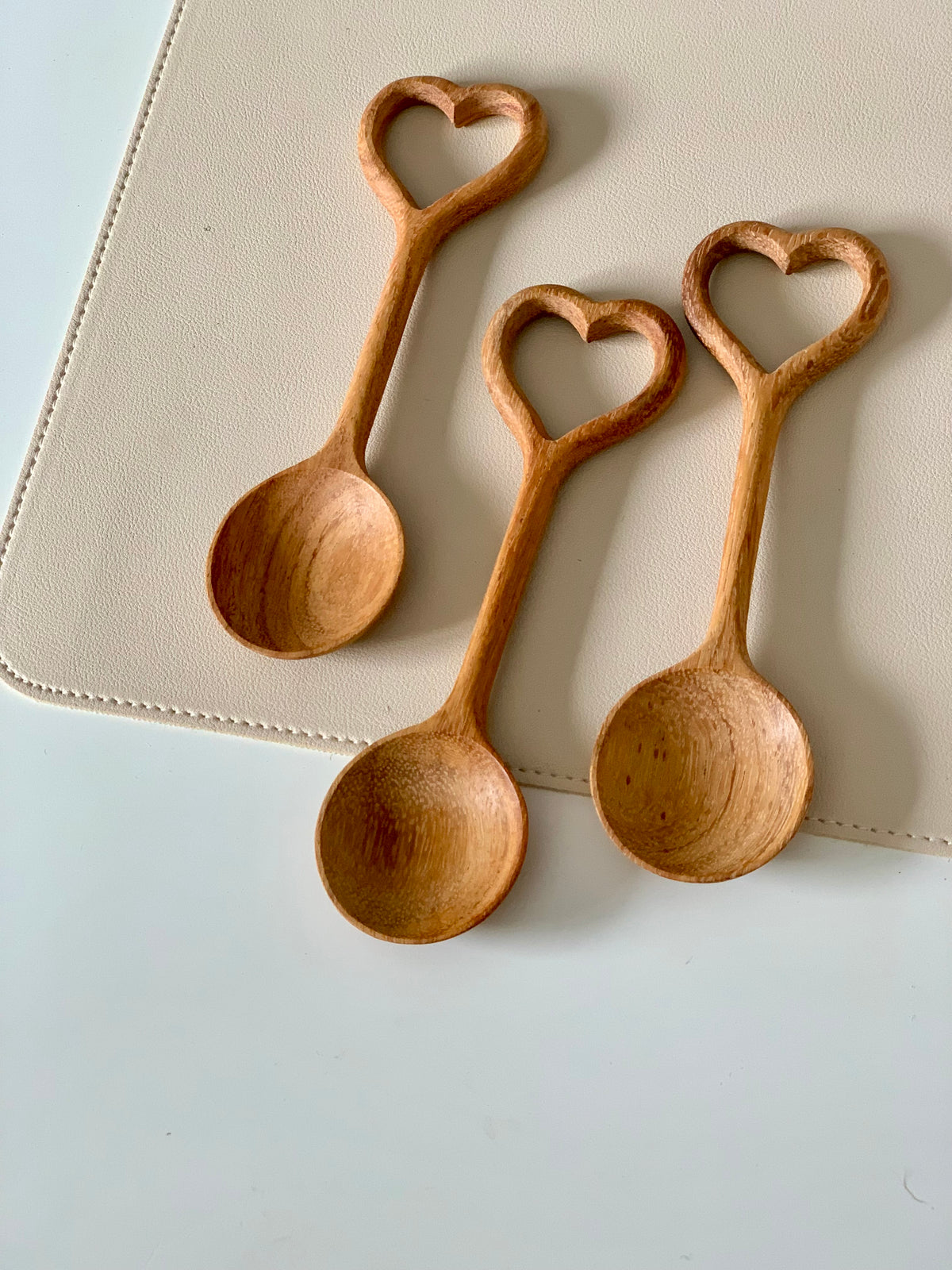 Three wooden spoons with heart-shaped handles resting on a light beige placemat on a white surface