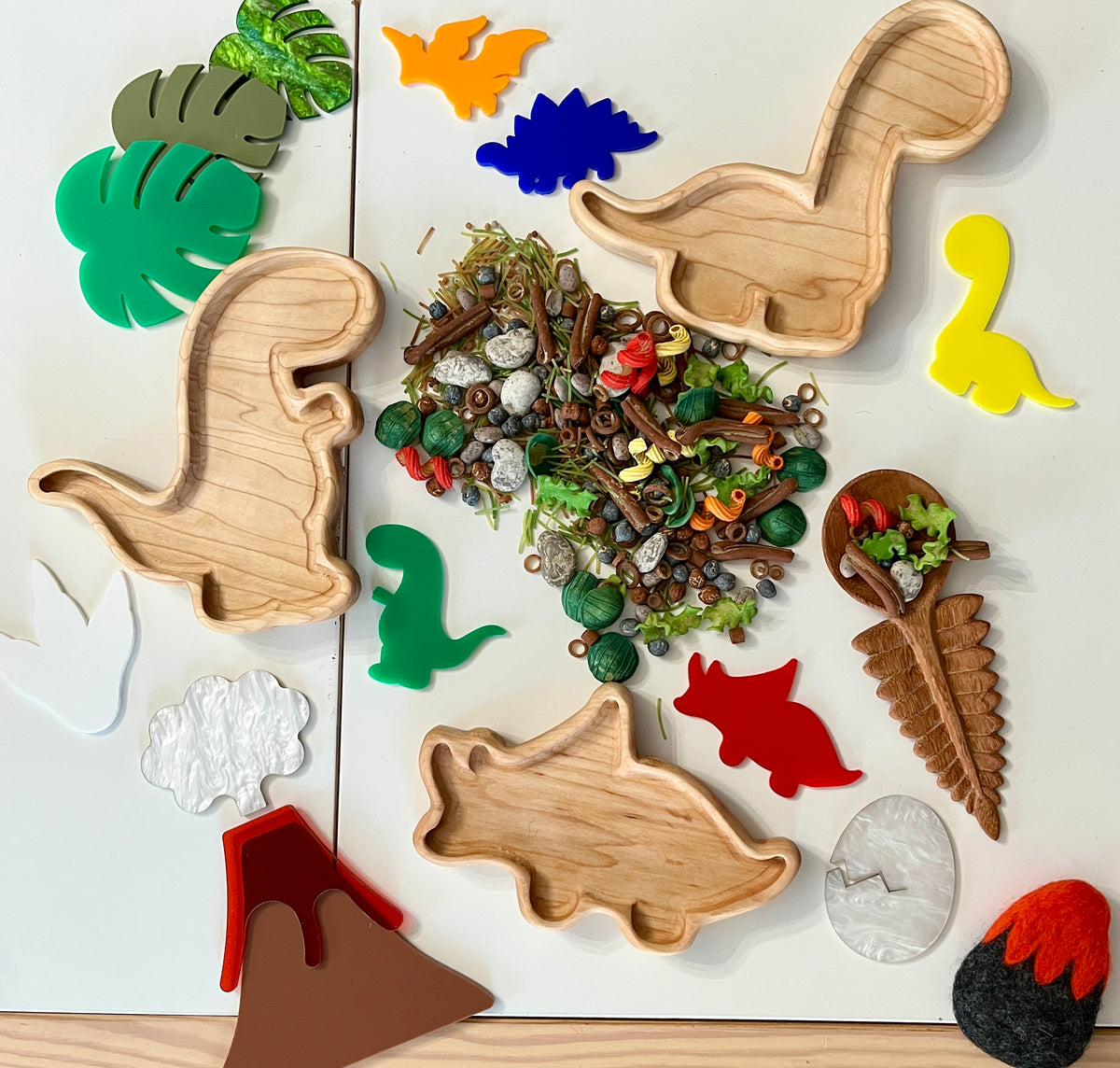 Wooden dinosaur-shaped trays with colorful dinosaur cutouts, green leaves, natural play materials, and a brown cone-shaped toy on white surface