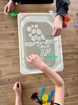 White tray with a stone path and fence design, held by children placing colorful translucent acrylic pegs shaped like plants and garden elements, featuring STEM Insert – Acrylic Insert with Different Peg Options - Neighborhood, Veggie Garden, Flower Garden, Under the Sea + More, handcrafted, homeschool, education resources.