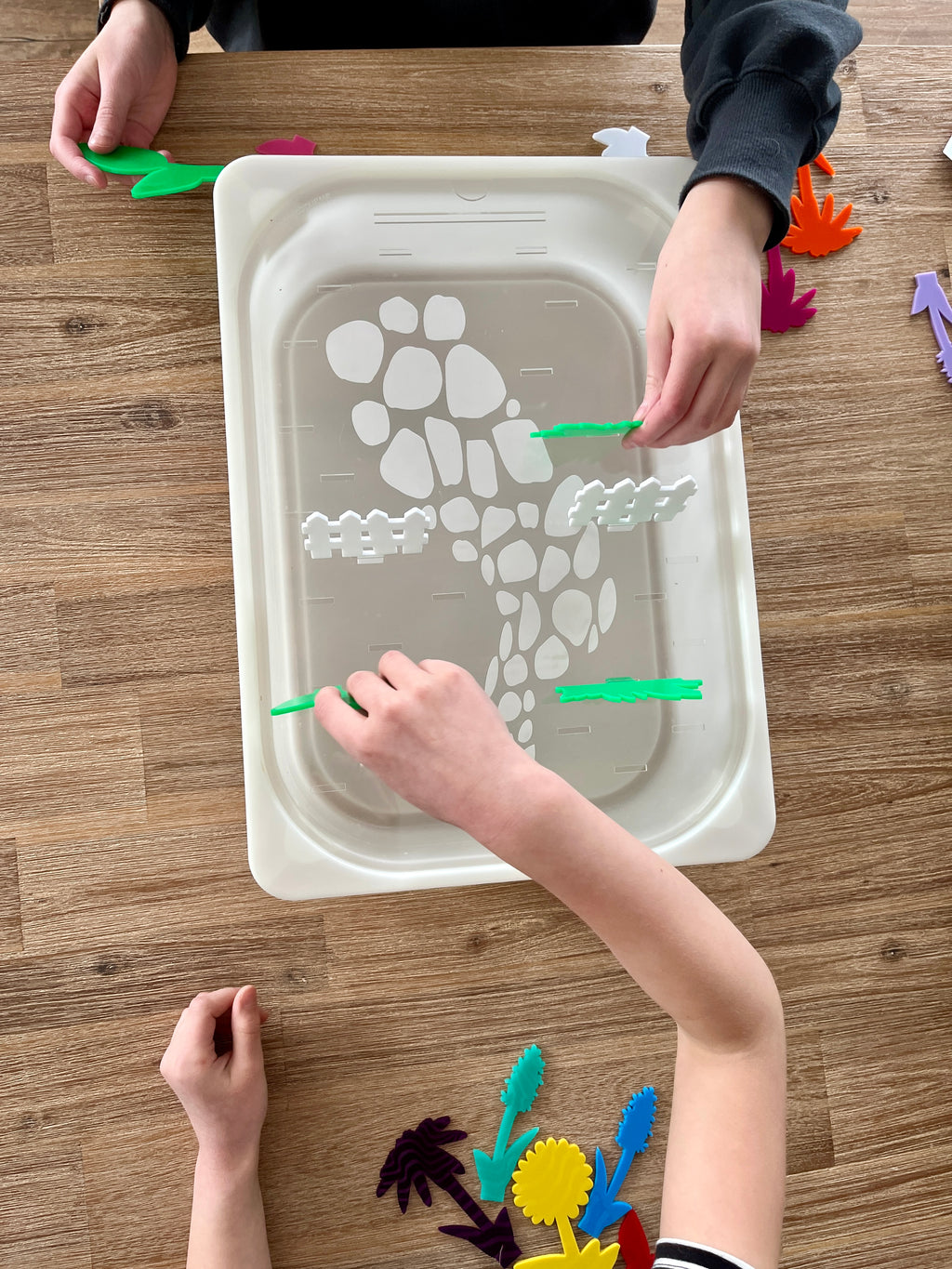 White tray with a stone path and fence design, held by children placing colorful translucent acrylic pegs shaped like plants and garden elements, featuring STEM Insert – Acrylic Insert with Different Peg Options - Neighborhood, Veggie Garden, Flower Garden, Under the Sea + More, handcrafted, homeschool, education resources.