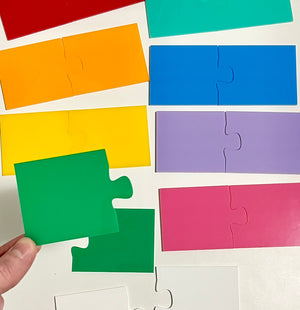 Colorful wooden puzzle pieces arranged on a white background with a hand holding a green piece, showcasing Little Dry Erasables - 2 Part Puzzles, handcrafted homeschool education resources.