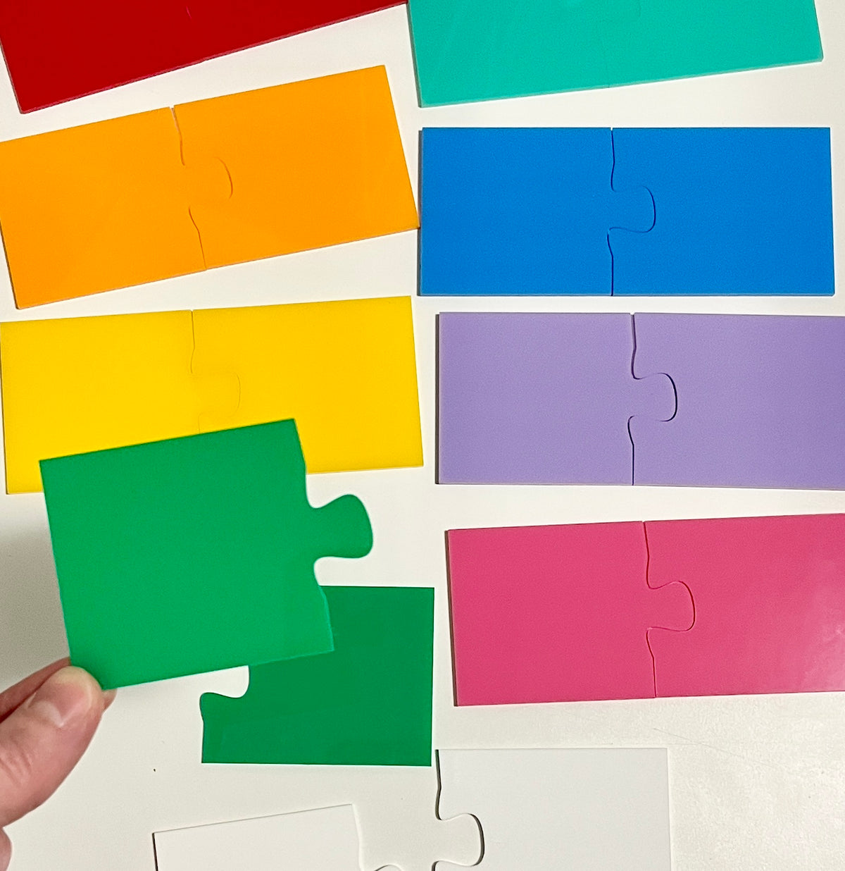 Colorful wooden puzzle pieces arranged on a white background with a hand holding a green piece, showcasing Little Dry Erasables - 2 Part Puzzles, handcrafted homeschool education resources.